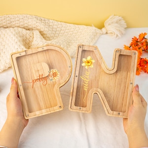 May include: Two wooden money boxes shaped like the letters "P" and "N". The "P" has the word "petty" written on it in cursive and the "N" has the word "midlife" written on it in cursive. Both money boxes have a slot on the top for coins and a clear plastic window on the front.