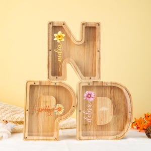 May include: Wooden letter money boxes with clear plastic windows. The letters spell "NPD" and each letter has a different floral design and a name on it. The names are "Nidlia", "Petty", and "Debra".