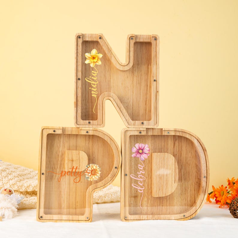 May include: Wooden letter money boxes with clear plastic windows. The letters spell "NPD" and each letter has a different floral design and a name on it. The names are "Nidlia", "Petty", and "Debra".