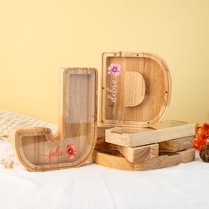 May include: Two wooden letter money boxes, one shaped like a "J" and the other shaped like a "D". The "J" is decorated with a red poppy and the name "julie" in cursive. The "D" is decorated with a pink flower and the name "debra" in cursive.