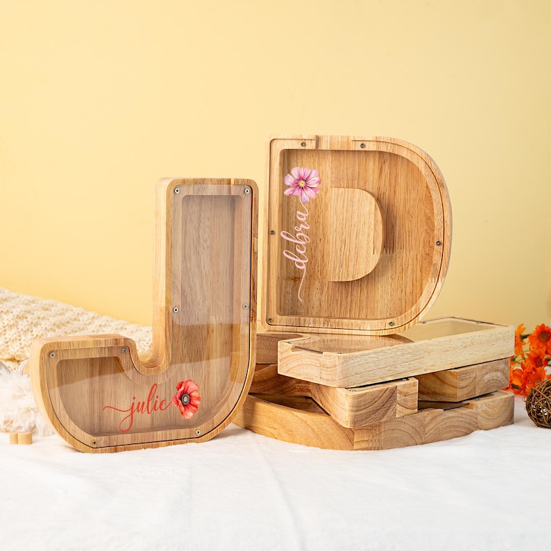 May include: Two wooden letter money boxes, one shaped like a "J" and the other shaped like a "D". The "J" is decorated with a red poppy and the name "julie" in cursive. The "D" is decorated with a pink flower and the name "debra" in cursive.