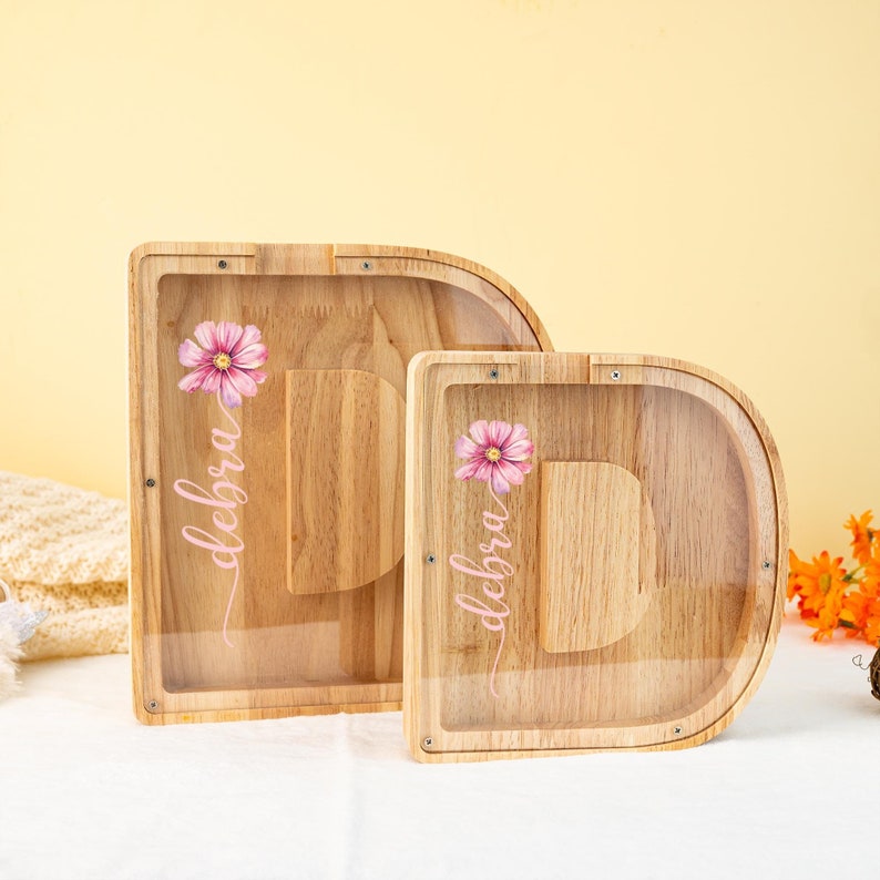 May include: Two wooden money boxes shaped like the letter D. Both money boxes have a clear plastic front and a slot on the top. The money boxes are decorated with a pink flower and the name "Debra" written in pink cursive.
