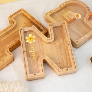 May include: Wooden letter N with a clear acrylic insert, featuring a yellow daffodil and the name "Midia" written in cursive. The letter is a decorative box with a slot for coins or other small items.