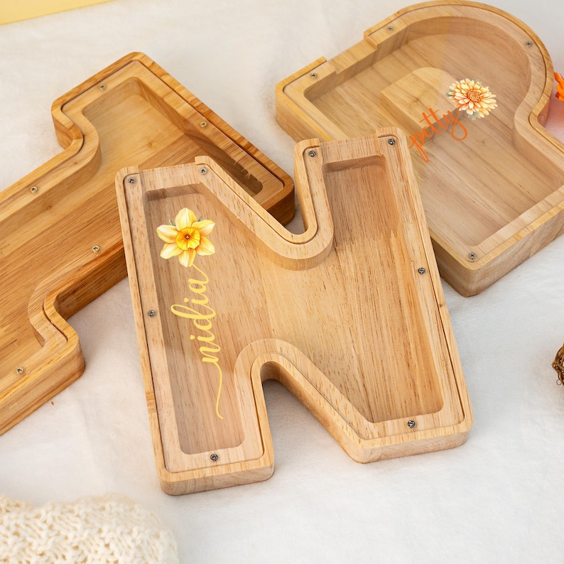 May include: Wooden letter N with a clear acrylic insert, featuring a yellow daffodil and the name "Midia" written in cursive. The letter is a decorative box with a slot for coins or other small items.