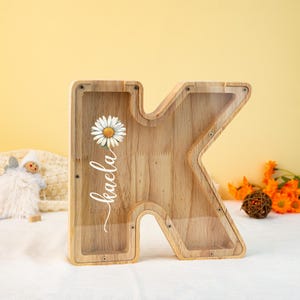 May include: Wooden letter K money box with a clear front panel. The front panel features a white daisy and the name "Kaela" written in cursive.