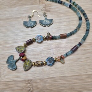 May include: A necklace and earrings set featuring ginkgo leaf charms and colorful beads. The necklace has a mix of blue, green, brown, and red beads, with a floral design on some of the beads. The earrings are simple ginkgo leaf charms.