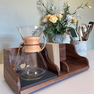 May include: A wooden coffee station with a glass pour-over coffee maker, a stack of filters, and a gray mat. The station also features a decorative floral arrangement and utensils in a rustic container. The wood has a rich, dark brown finish.