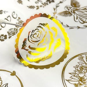 Peut inclure: Gros plan de plusieurs autocollants ronds avec des accents dorés. L'un des autocollants présente un motif de rose dorée, tandis que d'autres affichent des motifs floraux et d'anges. Les autocollants ont un bord festonné et sont sur fond blanc.
