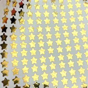 May include: Sheet of gold star stickers. The stickers are arranged in rows and columns. Some of the stars are a shiny gold color, while others are a darker gold or bronze color. The background is a light gray color.
