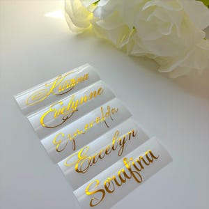 May include: White name tags with gold cursive lettering. The names Kristina, Evelynne, Esmeralda, Evelyn, and Serafina are displayed. A white flower is in the background.