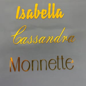 Könnte beinhalten: Drei goldfarbene Namensausstiche: "Isabella", "Cassandra" und "Monnette" werden vor einem grauen Hintergrund angezeigt. Die Namen sind in einer Schreibschrift, mit einem reflektierenden, metallischen Finish. Die Namen sind wahrscheinlich für eine Party oder Veranstaltung.