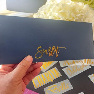 以下が含まれることがあります： 金色の筆記体で「Scarlet」と書かれた濃い青色の封筒。他の金色の名前デカールが下にいくつか見えます。背景には白い花と灰色の表面があります。