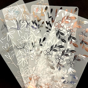 May include: A collection of clear, translucent sheets featuring floral designs in various metallic finishes, including silver, gold, and rose gold. The designs include flowers, leaves, and stems, creating an elegant and botanical theme.