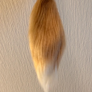 Könnte beinhalten: Ein Tier-Schwanz-Accessoire aus Kunstfell mit einem Farbverlauf, der von einem warmen Goldbraun an der Oberseite zu einer leuchtend weißen Spitze übergeht. Der Schwanz ist an einem kleinen schwarzen Metall-Karabinerhaken befestigt.