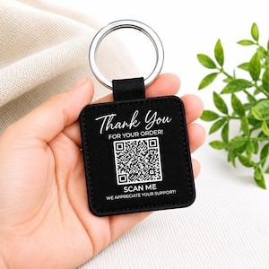 May include: A square black keychain featuring white text that reads Thank You FOR YOUR ORDER! with a central QR code and the text SCAN ME WE APPRECIATE YOUR SUPPORT! below it. The keychain has a silver metal ring attached by a black fabric loop.