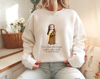 Puede incluir: Sudadera blanca con una ilustración de una santa sosteniendo flores. La santa lleva un hábito marrón y una capa amarilla. El texto en la sudadera dice: "No nos cansemos de orar, la confianza hace milagros, Santa Teresa de Lisieux."