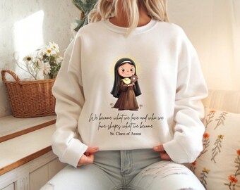 May include: White sweatshirt featuring an illustration of St. Clare of Assisi holding a flower. The design includes the quote "We become what we love and who we love shapes what we become" and the saint's name.