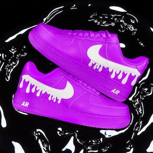 May include: Bright purple trainers with white Nike swooshes and dripping paint effect. The word "AIR" is printed on the side. The shoes are set against a black background with white liquid patterns.
