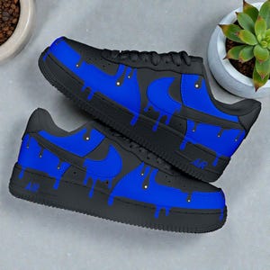 Può includere: Un paio di sneakers nere e blu con un design a goccia unico. Le scarpe presentano una base nera con dettagli blu, tra cui l'iconico logo swoosh e un effetto vernice gocciolante. La parola "AIR" è visibile sul lato della scarpa.