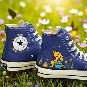 Puede incluir: Zapatillas altas azul marino con cordones y suelas blancas. Las zapatillas presentan diseños bordados de Winnie the Pooh, abejas y detalles florales. El logotipo de Converse All Star es visible en el lateral.