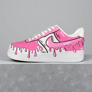 以下が含まれることがあります： ピンクと白のアスレチックシューズ、ユニークなドリップデザイン。 ピンクのアッパーに白いスウッシュと靴紐が付いています。 黒いアウトラインがドリップ効果とデザイン要素を定義しています。 ソールは白です。