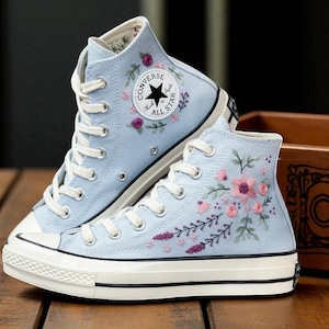 Puede incluir: Un par de zapatillas altas de color azul claro con cordones y suelas blancas. Las zapatillas presentan diseños florales bordados en rosa, morado y verde. El logotipo de Converse es visible.