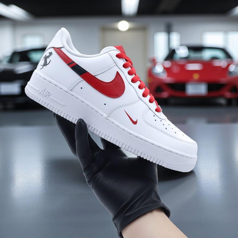 May include: White leather sneaker with red accents, including the Nike swoosh, laces, and a stripe. Features a low-top design with the word "AIR" on the side. A small logo is visible near the heel. The shoe is held by a gloved hand.