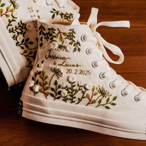 May include: Cream-colored high-top sneakers with intricate floral embroidery in green, white, and red. The text "Jessica & Lucas 20.2.2025" is embroidered on the side. The sneakers have white laces and metal eyelets.