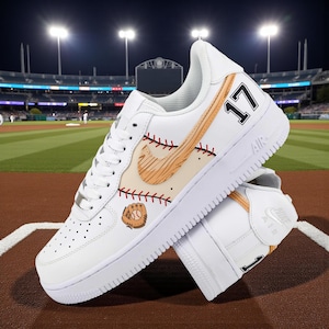 Pode incluir: Tênis de couro branco com designs temáticos de beisebol. Apresentam uma luva de beisebol, costuras vermelhas e o número 17. Os tênis estão em um fundo de campo de beisebol.