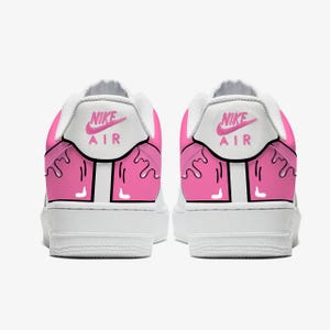以下が含まれることがあります： ピンクのアクセントが付いた白いナイキエアフォース1スニーカーのペア。かかとには、ピンクのナイキエアロゴが付いた白いパネルがあります。ピンクのペイントのようなデザインが靴の背面を滴り落ちています。靴は白い背景にあります。