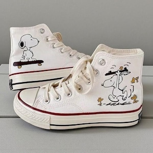 以下が含まれることがあります： スヌーピーのイラストが描かれた白いハイカットキャンバススニーカーのペア。片方の靴はスケートボードに乗ったスヌーピー、もう片方はトレイを持ったスヌーピーが描かれています。靴は赤と黒のアクセントと白い靴紐が付いています。