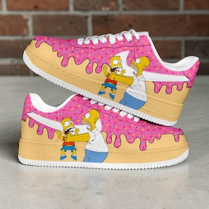Pode incluir: Tênis rosa e bege personalizados com um design de donut e personagens de desenhos animados. Os sapatos apresentam cadarços e solas brancas, com um padrão de respingos coloridos na parte superior rosa. Os personagens são Homer e Bart Simpson.
