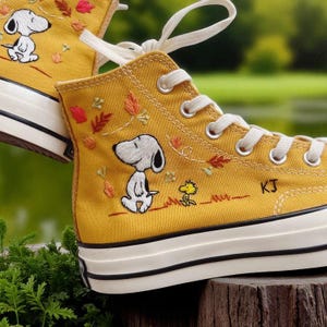 May include: Mustard yellow high-top sneakers featuring embroidered Snoopy and Woodstock designs. The canvas shoes have white laces, a white rubber sole, and a black stripe. The sneakers have the initials "KJ" embroidered on the side.