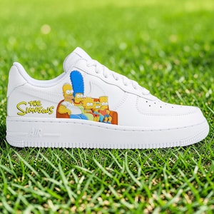 Puede incluir: Zapatilla deportiva blanca con una colorida ilustración de la familia Simpson en el lateral. La zapatilla tiene la palabra "Simpsons" en amarillo y la palabra "AIR" en la suela. La zapatilla está sobre un fondo de hierba verde.