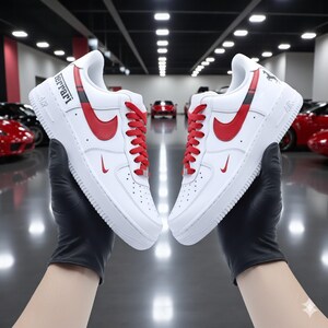 May include: Pair of white sneakers with red accents, including the Nike swoosh and laces. The word "Ferrari" is printed on the side. The shoes are held up by a person wearing black gloves.