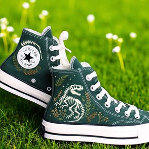 Puede incluir: Un par de zapatillas altas verde oscuro con cordones y suelas blancas. Las zapatillas presentan un gráfico de esqueleto de dinosaurio blanco y el logotipo de Converse All Star. Se encuentran sobre un fondo de hierba verde y flores blancas.