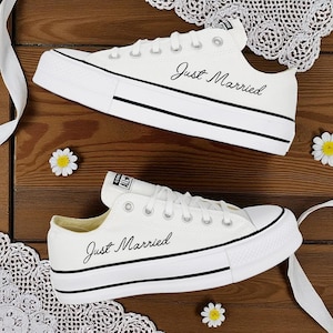 Puede incluir: Dos zapatillas de lona blancas con ribetes negros y la inscripción "Just Married". Están sobre una superficie de madera con encaje blanco y pequeñas margaritas.