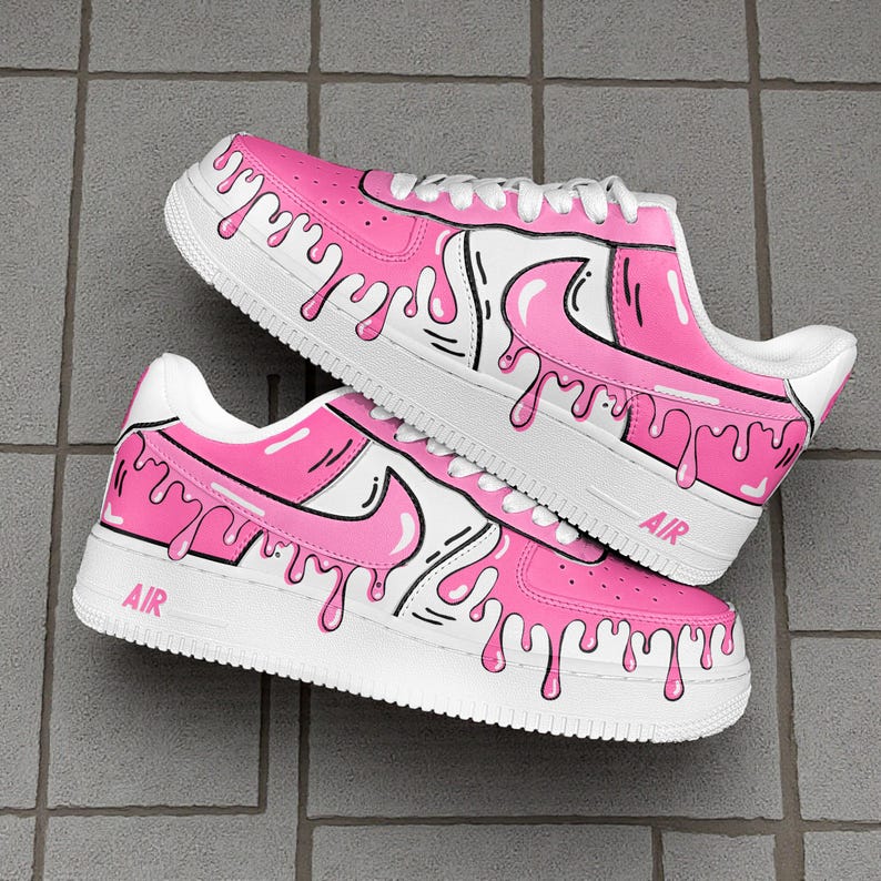 以下が含まれることがあります： ピンクと白のカスタムスニーカーのペア。ドリップデザインが施されています。靴は白いソールと靴紐で、側面に「AIR」の文字がプリントされています。ピンクの塗料が靴の側面を伝って滴り落ちているように見えます。
