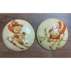 May include: Two vintage, round wall decorations featuring a cartoon bear skiing. The bear wears a red hat, scarf, and mittens. The background is a cream color, and the skis are brown. The decorations are on a wooden surface.