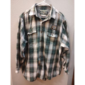 May include: Green Bay Packers flannel shirt with a plaid pattern in green, white, and tan. The shirt has two chest pockets and a Packers logo patch. The shirt is button-down with a collar and long sleeves.