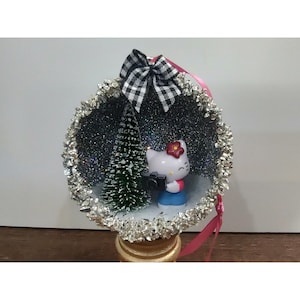 Könnte beinhalten: Eine kugelförmige Weihnachtsdekoration mit einer weißen Hello-Kitty-Figur, die eine schwarze Kamera neben einem kleinen grünen Tannenbaum hält. Das Innere ist dunkel mit silbernem Glitzer und der äußere Rand ist mit Silberfolie verziert. Eine schwarz-weiß karierte Schleife sitzt oben mit einem rosa Band.