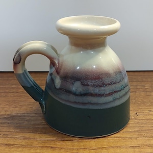 Puede incluir: Una taza de cerámica con un esmalte verde, blanco y morado. La taza tiene una base ancha y redondeada y un borde acampanado. El asa está unida al lado de la taza.