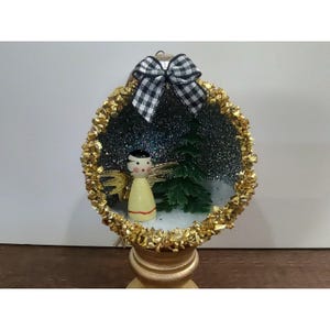 Puede incluir: Un adorno redondo y hueco con un borde dorado texturizado y un lazo de cuadros vichy en blanco y negro en la parte superior. Dentro, una pequeña figura de ángel de madera con alas doradas junto a un pino verde sobre un fondo oscuro, brillante y estrellado.