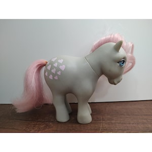 May include: A gray toy pony with pink hair and tail. The pony has blue eyes and pink heart-shaped designs on its side. The toy is standing on a wooden surface.