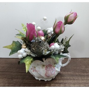 May include: A decorative arrangement in a white porcelain teacup with pink floral patterns. The centerpiece features dark pink rosebuds, green leaves, white berries, gold spheres, and a glittery dark gray bird figure nestled among the foliage.