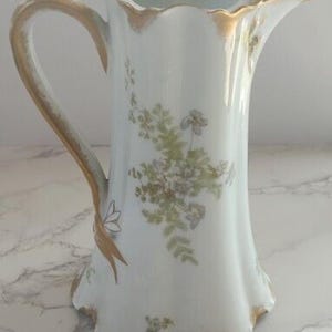 Puede incluir: Una jarra de crema de porcelana blanca con un borde dorado y un diseño floral. La jarra tiene una base acanalada y un asa.