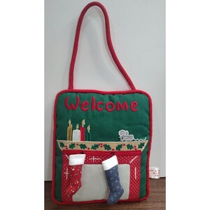 May include: A rectangular green fabric wall hanging with a red border and a red hanging loop. It features the embroidered red text Welcome, a fireplace scene with four candles, a small mouse, and two miniature stockings with white cuffs.