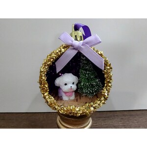 以下が含まれることがあります： ピンクのスカーフを巻いた白い犬のフィギュアと小さな緑の松の木が入った丸いオーナメント。フレームはゴールドのグリッターで装飾され、上部には薄紫色のリボン結びと、吊り下げ用の紫色のループが付いています。