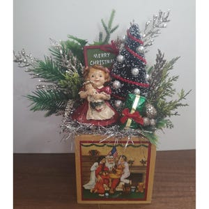 Könnte beinhalten: Ein Weihnachtsgesteck mit einer Mädchenfigur im roten Kleid, die eine Puppe hält, einem kleinen Tannenbaum mit weißen Perlen und einem grünen Geschenk. Ein Schild trägt die Aufschrift MERRY CHRISTMAS. Der Sockel ist ein Holzblock mit Weihnachtsmann-Motiv.