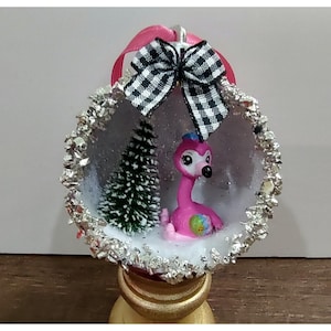 Puede incluir: Adorno navideño decorativo con una figura de flamenco rosa dentro de un globo transparente lleno de nieve. El adorno está adornado con purpurina plateada y un lazo a cuadros blanco y negro. También hay un pequeño árbol de Navidad verde en el interior.